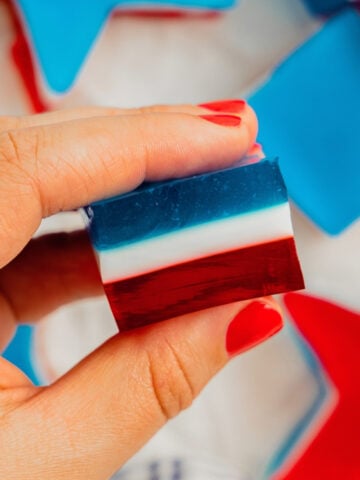 A woman's hand with red painted nails holds a red, white, and blue layered jello shot.