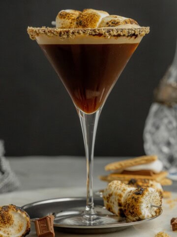 A martini glass rimmed with crushed graham crackers is filled with a s'mores espresso martini and garnished with a toasted marshmallow skewer.