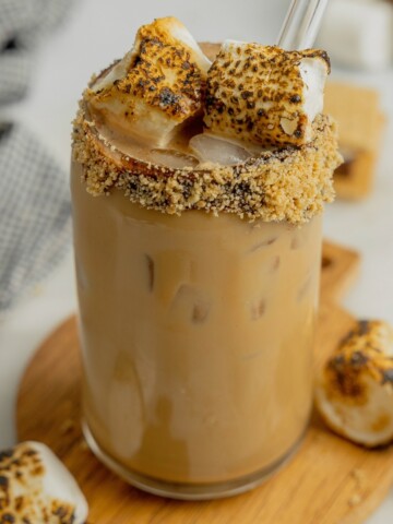 A tall glass rimmed with chocolate sauce and crushed graham crackers is filled with a s'mores mocha latte and garnished with toasted marshmallows.