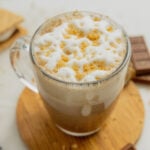 A clear glass mug filled with a campfire mocha sits on a small wooden coaster surrounded by s'mores and pieces of chocolate.
