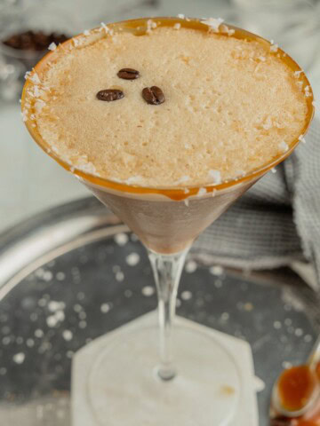 A classic martini glass is rimmed with caramel sauce and flaky sea salt and holds a salted caramel espresso martini that is garnished with three coffee beans. The glass sits on a white marble coaster.