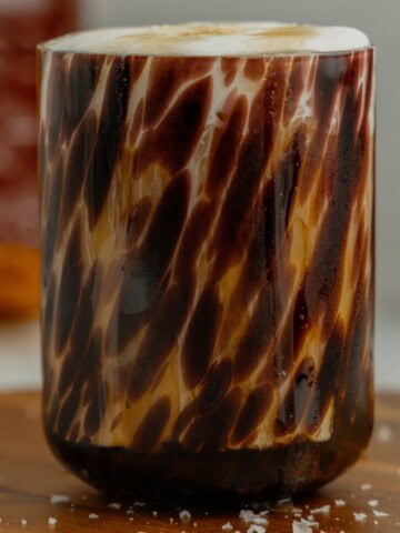 A tortoise shell glass filled with salted maple cold foam cold brew coffee sits on a wooden butting board with flaky sea salt sprinkled around it.