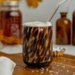A tortoise shell glass filled with salted maple cold foam cold brew coffee sits on a wooden butting board with flaky sea salt sprinkled around it and a clear glass straw in the glass.