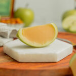 A caramel apple jello shot served in a granny smith wedge sits on a white marble coaster on a wooden serving board.