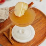 An apple cider martini in a coupe glass garnished with fresh apple slices and a cinnamon stick sits on a marble coaster that is set on a wooden board.
