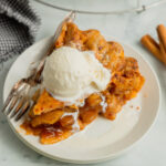 A slice of apple pie in a cheddar cheese crust is topped with a scoop of vanilla ice cream and sitting on a white dessert plate.