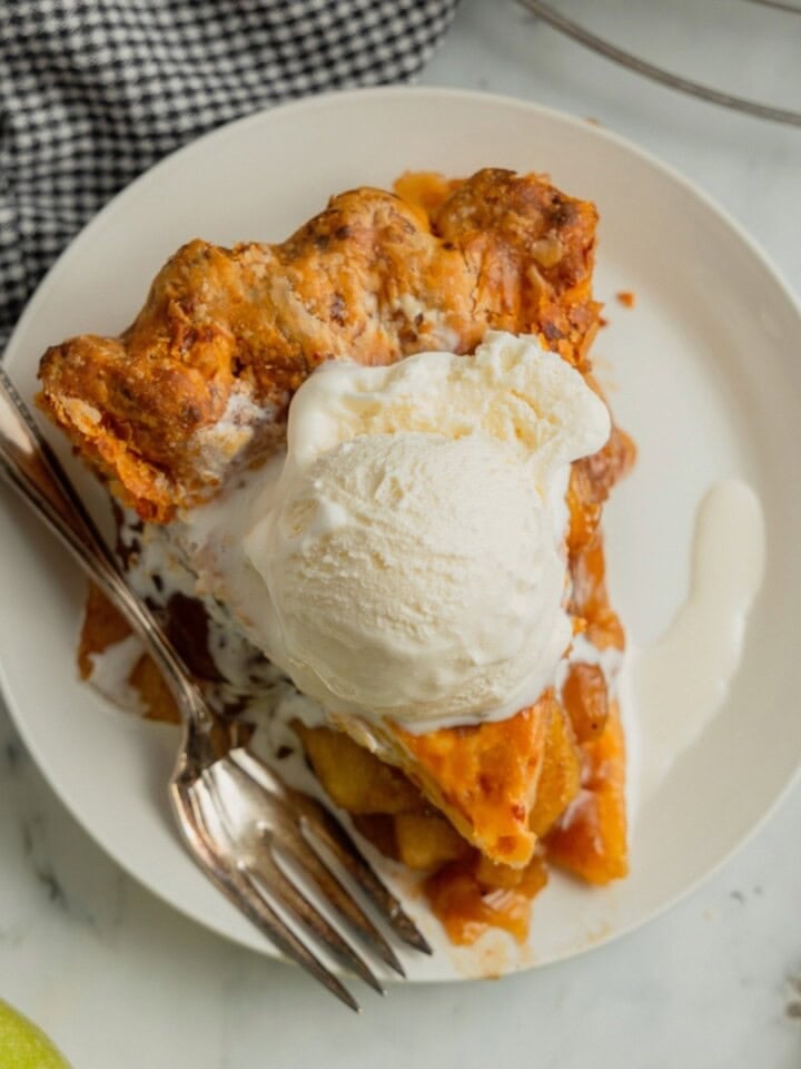 An overhead view of a slice of Wisconsin apple pie with cheddar cheese crust, topped with a scoop of vanilla ice cream, on a white dessert plate with a fork resting beside it.