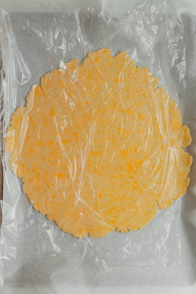 A batch of cheddar cheese pie crust rolled out and covered with plastic wrap.