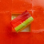 A square of a watermelon jello shot tipped upright in the pan of shots to show the dark green, light green, and deep pink layers.